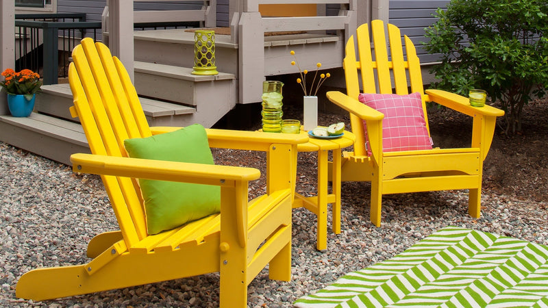 Two POLYWOOD Classic Folding Adirondack Chairs in Lemon are arranged on a gravel patio with a patterned rug and pillows.