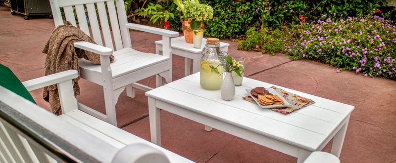 An assortment of white POLYWOOD furniture sits on a concrete patio next to a blooming flower garden.