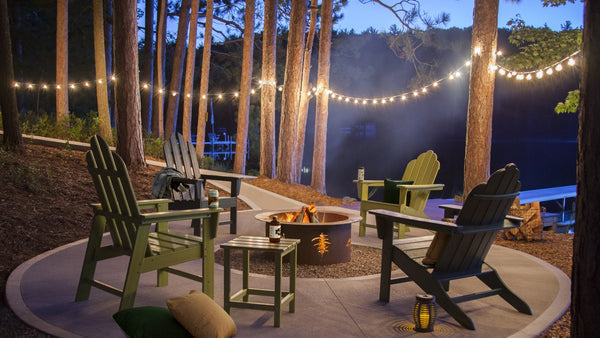 Assorted green POLYWOOD Long Island Adirondack chairs and side tables circle a fire pit in a wooded area by a lake.