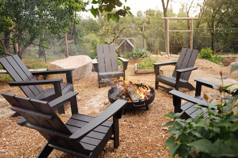 Black POLYWOOD Modern Adirondack Chairs sit around a fire pit in a mulched area with trees and a vegetable garden in the background.