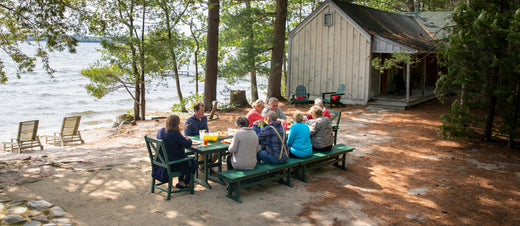 People sit on POLYWOOD Braxton Dining Arm Chairs and Farmhouse Trestle 65 Inch Benches around Farmhouse 37 Inch by 72 Inch Dining Tables in Green by a lakeside cabin in the woods.