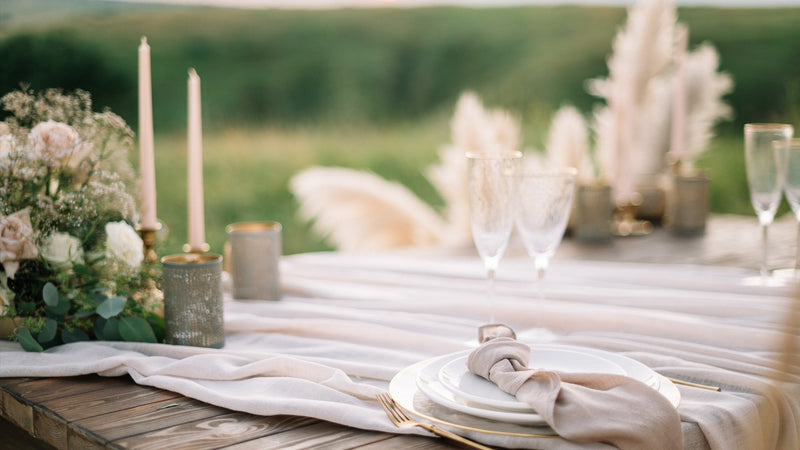 A wooden table is set for an elegant outdoor event with a linen tablecloth, candles, champagne flutes, flowers, plates, and gold flatware.