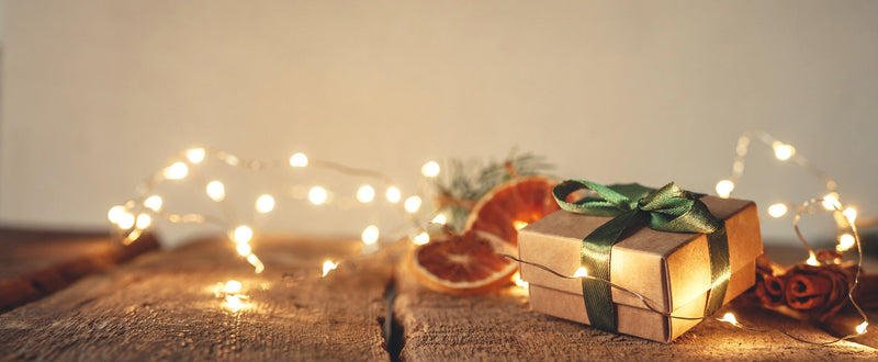 A small box tied with green ribbon sits on a wood surface with glowing fairy lights, dried fruit slices, and cinnamon sticks.