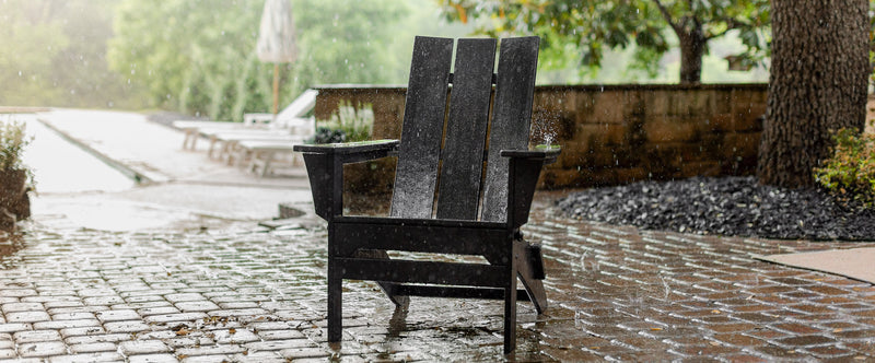 A POLYWOOD Modern Folding Adirondack Chair in Black stands in the rain on a brick patio.