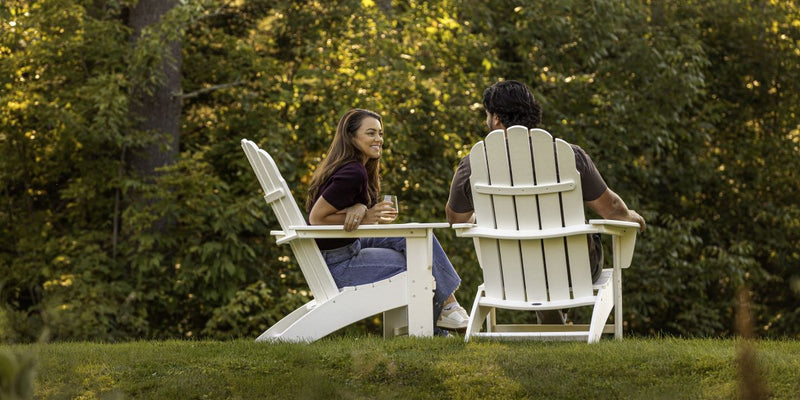Couple relaxing in white Adirondack patio chairs with a built-in table on a grassy hill, enjoying a drink at sunset.