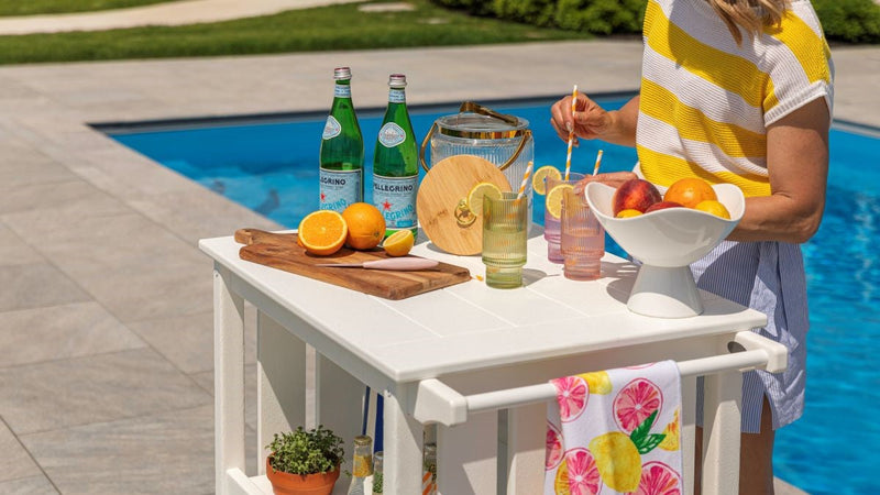 A woman in a yellow and white striped shirt mixes drinks at a white POLYWOOD coastal bar cart next to a swimming pool, showcasing an ideal outdoor home bar setup for entertaining.