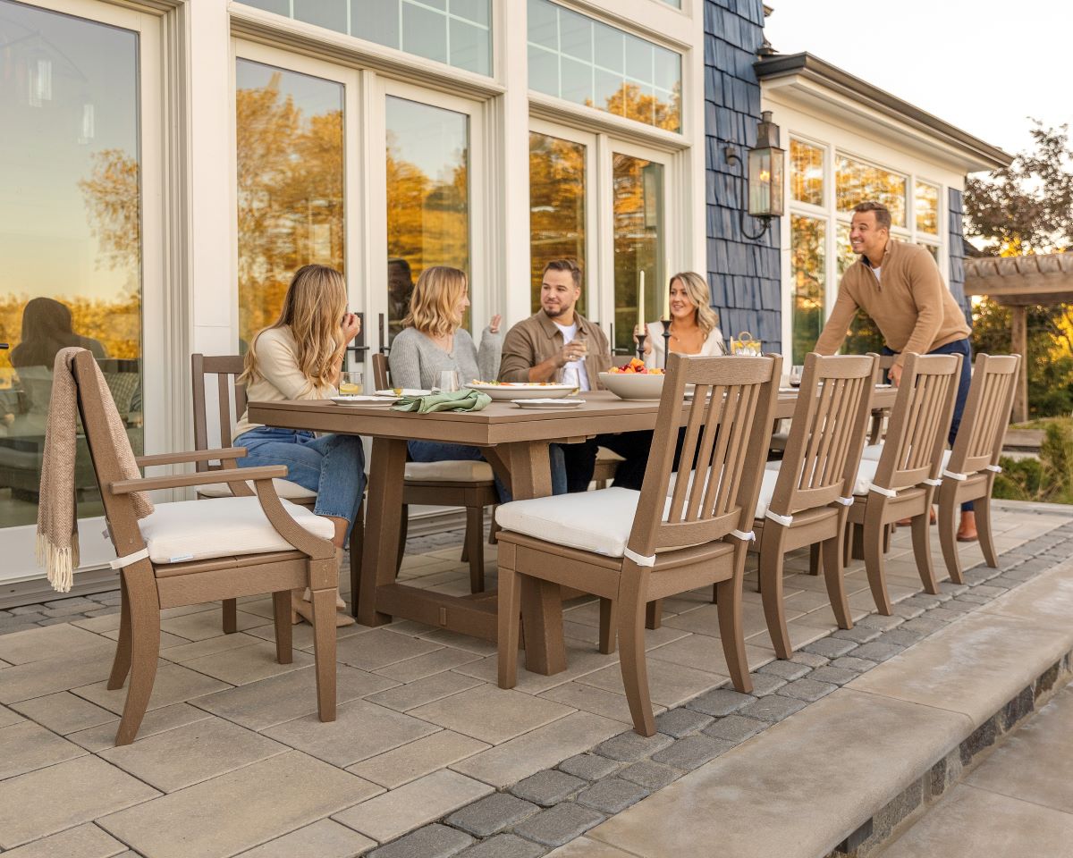 People chat at a tan PW Designer Series Estate 11 Piece A Frame Table Dining Set on a patio by a blue house with large windows.