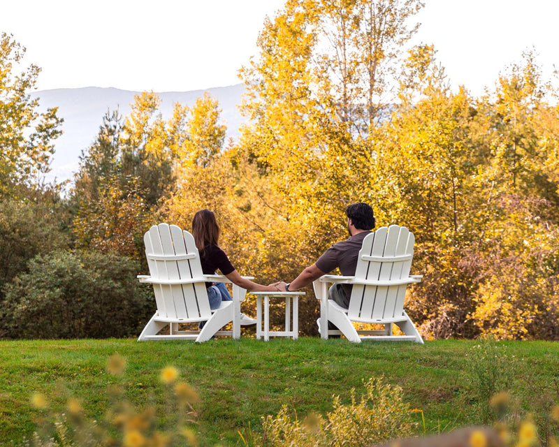 A couple holds hands while sitting at a POLYWOOD Vineyard 3 Piece Curveback Adirondack Set in White before a forest.