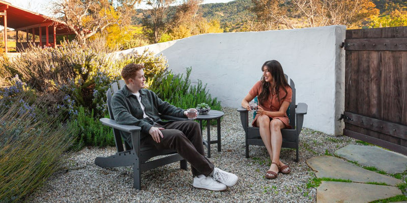 A couple sits in a grey POLYWOOD Classic Adirondack 3 Piece Set in a desert garden surrounded by plants and a white wall.