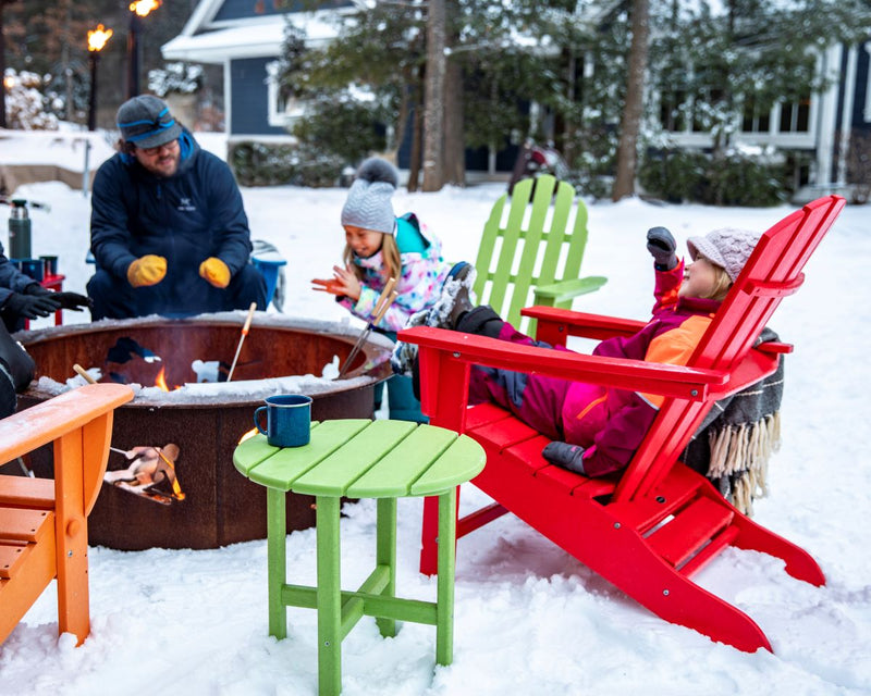 A family wearing coats, hats, and gloves gathers around a fire in colorful POLYWOOD Adirondack chairs on a snowy winter day.