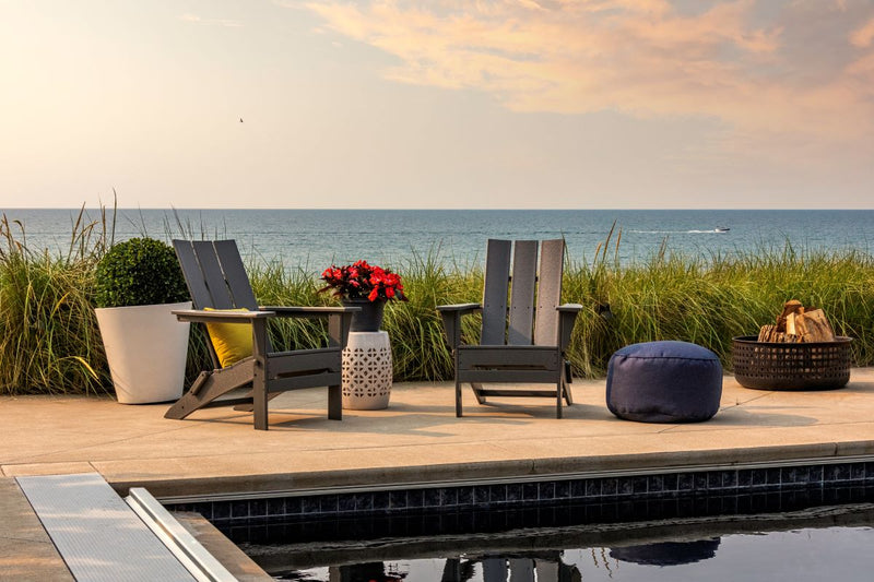 Grey POLYWOOD Folding Modern Adirondack Chairs sit on a poolside patio with grass and the ocean in the background.