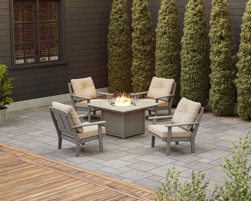 A grey POLYWOOD Mission 5 Piece Deep Seating Set with Fire Pit Table and beige cushions sits on a stone patio by tall hedges.