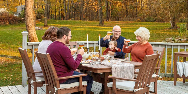 A family raises a toast around a brown POLYWOOD Vineyard 7 Piece Arm Chair Dining Set on a deck before a lawn and trees.