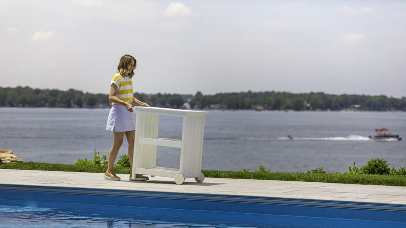 A woman pushes a white POLYWOOD portable outdoor bar cart with wheels on a poolside patio with a lake in the background.