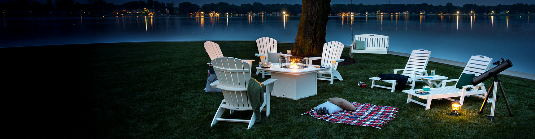 White POLYWOOD outdoor patio furniture from the Nautical Collection sits on a lawn by a large tree and a lake at night.