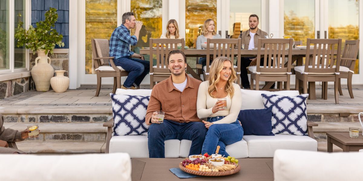 A man and a woman hold drinks and sit together on a tan PW Designer Series Estate Sofa with navy blue and white pillows.