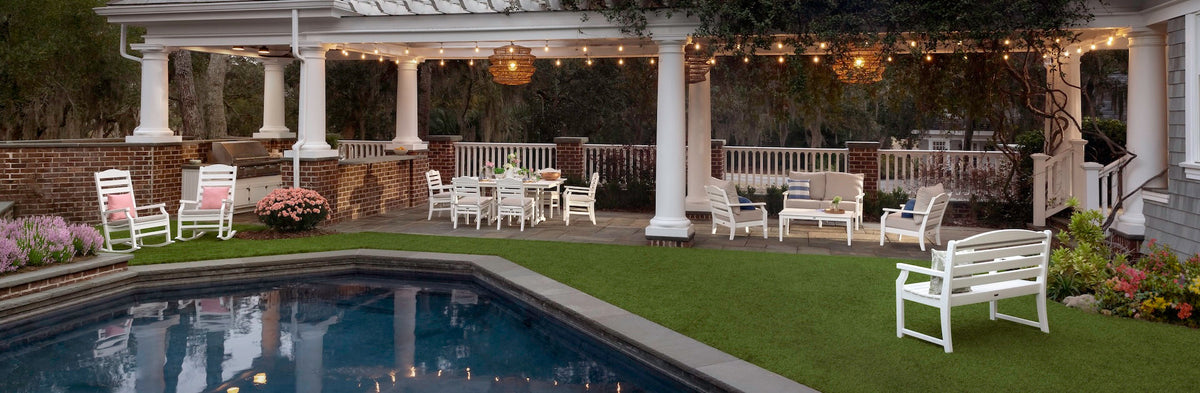 Various white furniture from the Draper James by POLYWOOD Savannah Collection sits in a backyard under a pergola and by a swimming pool.
