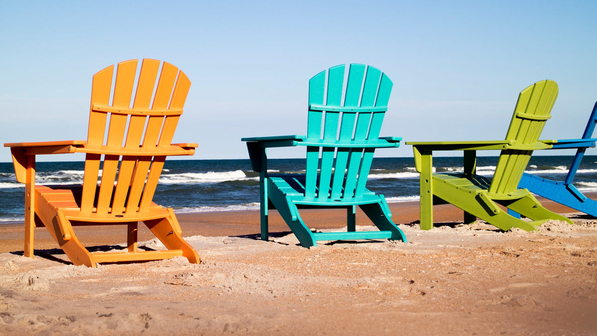 POLYWOOD Palm Coast Adirondack chairs in Tangerine, Aruba, Lime, and Pacific Blue face the ocean on a sandy beach.