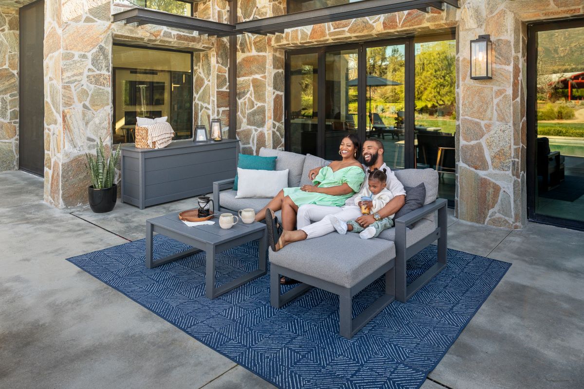 A family sits together on a grey POLYWOOD EDGE 4 Piece Modular Deep Seating Set with Ottoman on a patio behind a stone house.