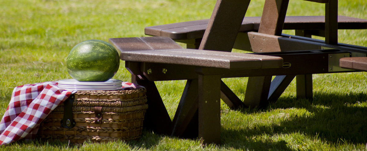 A brown POLYWOOD Park 53 Inch Octagon Table sits in a grassy area next to a basket holding plates, a table cloth, and a watermelon.