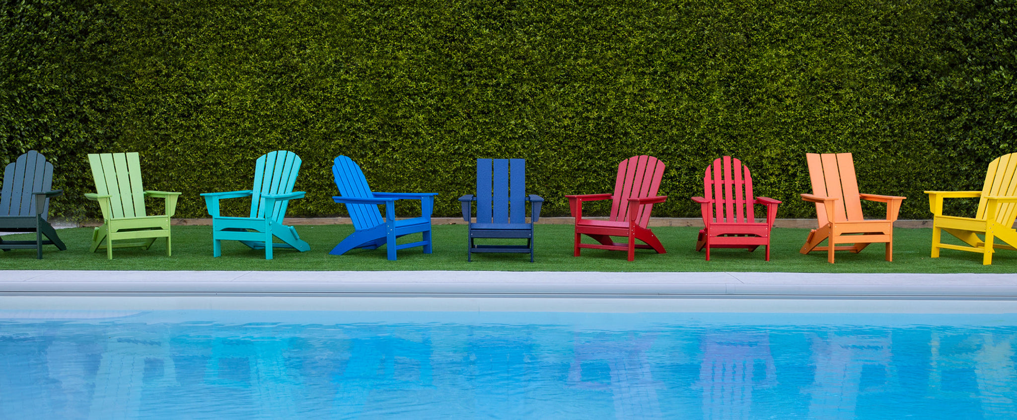 POLYWOOD Adirondack chairs in various styles and colors sit on a lawn by a swimming pool before a tall hedge.