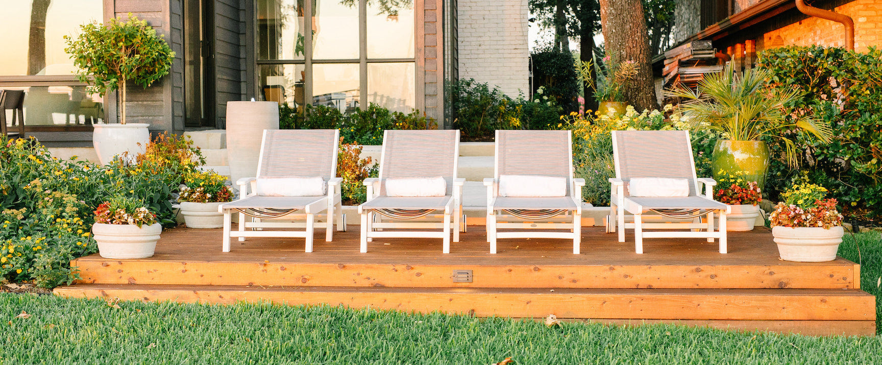 White POLYWOOD Coastal Chaises with Wheels and beige sling fabric sit on a wooden deck surrounded by landscaping.