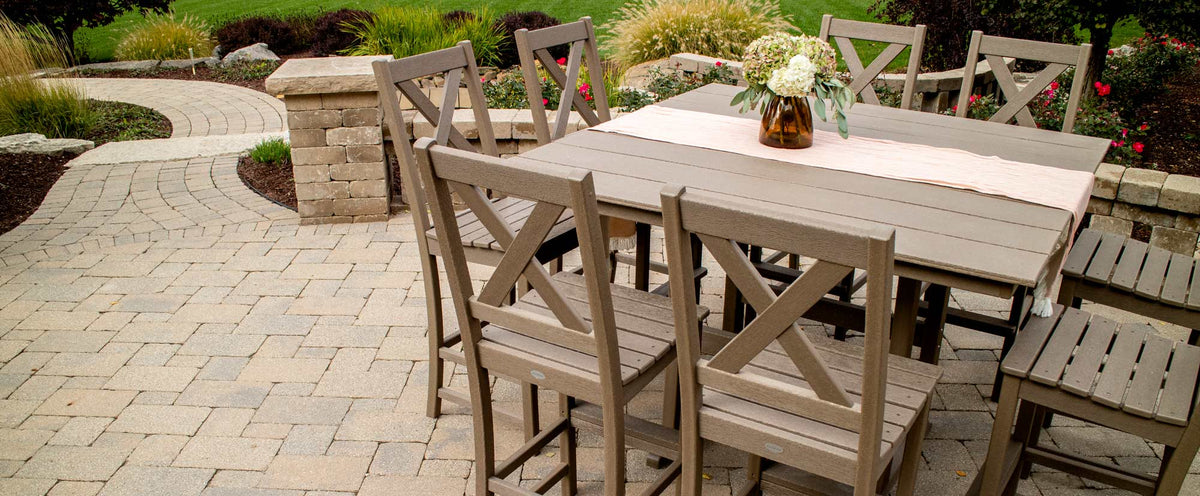 A brown POLYWOOD Braxton 9 Piece Farmhouse Trestle Bar Set with a beige runner and vase of flowers sits on a brick patio.