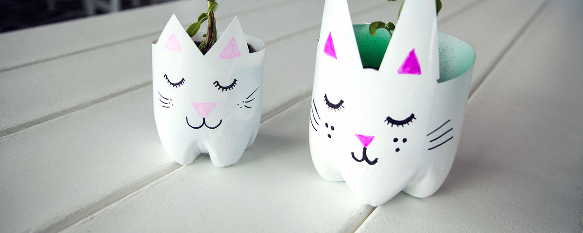 Two white cat shaped planters made from recycled bottles sit on a white slatted surface.