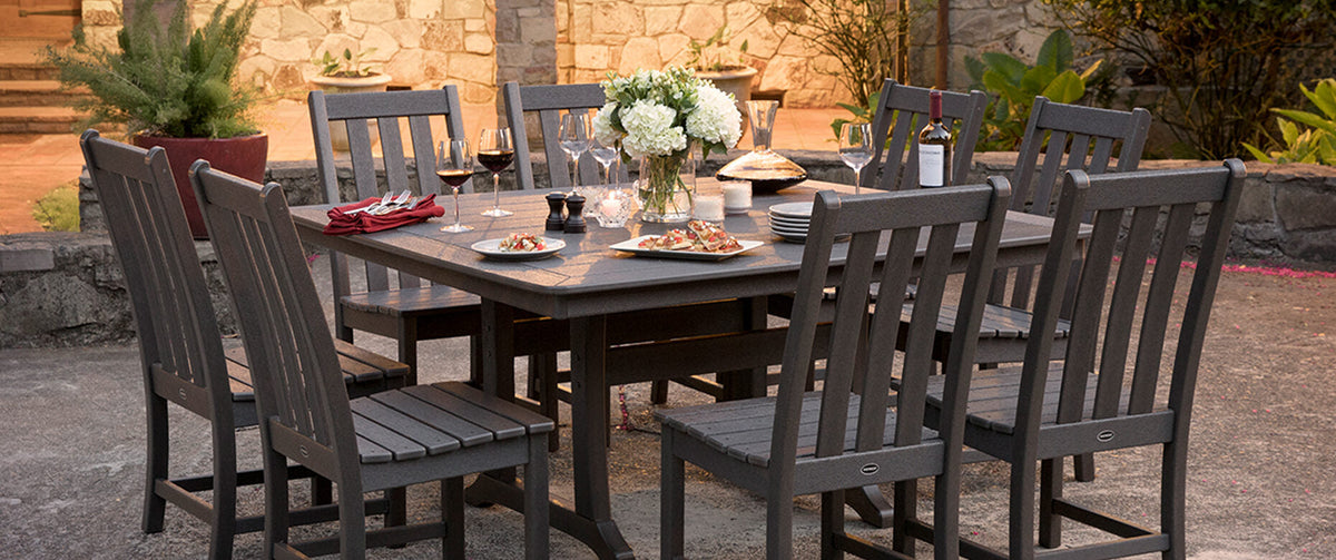 A POLYWOOD Vineyard 9 Piece Dining Set in Slate Grey is set for a meal on a concrete patio in front of a stone house.