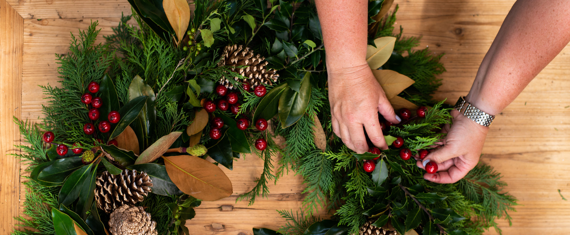 Hands adding red cranberries to a festive evergreen wreath decorated with pine cones and magnolia leaves.