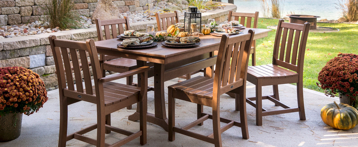 A dark brown POLYWOOD Traditional Garden 7 Piece Farmhouse Dining Set on a patio by a lake with pots of orange mums and fall decor surrounding it.
