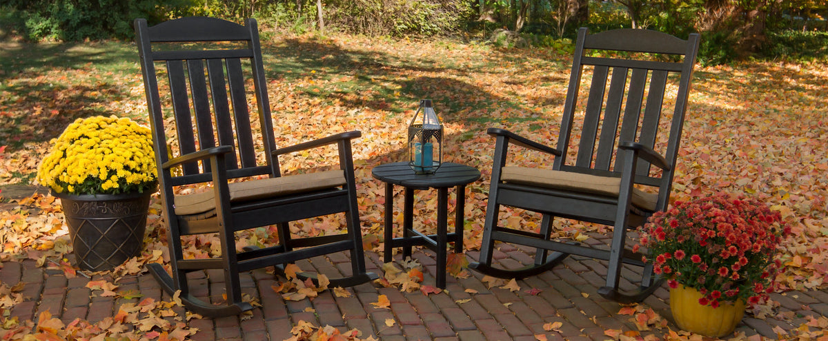 A black POLYWOOD Estate 3 Piece Rocking Chair Set and pots of mums sit on a brick patio by a lawn strewn with fallen leaves.