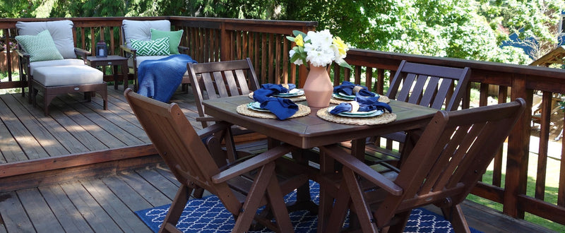 POLYWOOD Vineyard Folding Dining Chairs, a Nautical Trestle 37 Inch Dining Table, and a Newport 18 Inch End Table in Mahogany; and Vineyard Deep Seating Chairs and Vineyard Deep Seating Ottomans in Mahogany with Dune Burlap cushions sit on a deck.