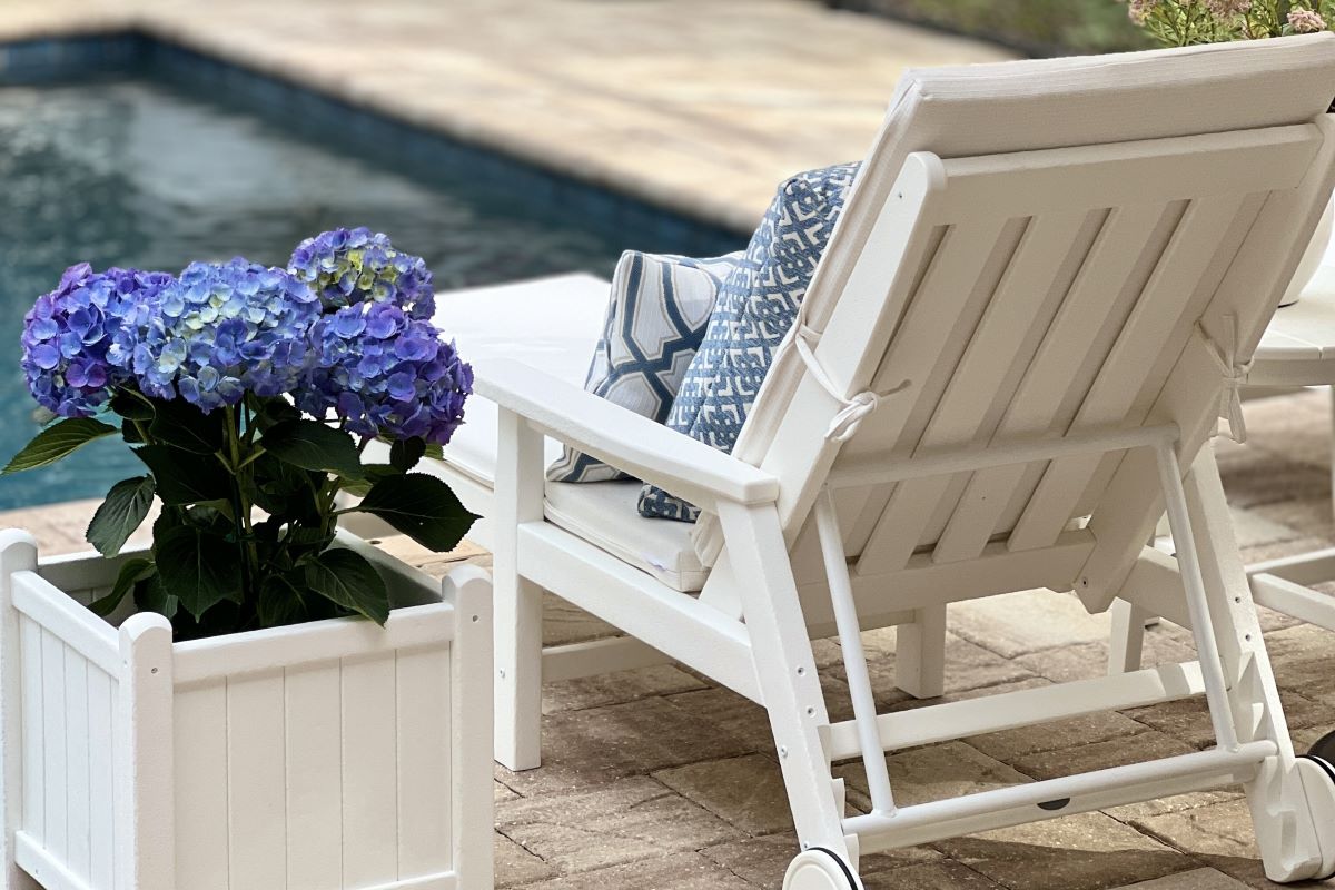 A white POLYWOOD planter box overflowing with vibrant purple hydrangeas sits beside a white chaise lounge next to a swimming pool, showcasing elegant outdoor planters.