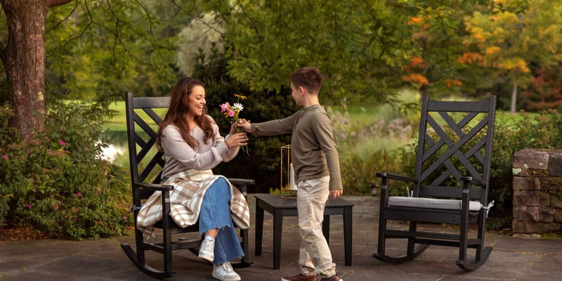 A boy hands flowers to a woman sitting in a black Chippendale 3 Piece Rocking Chair Set on a stone patio near tall trees.