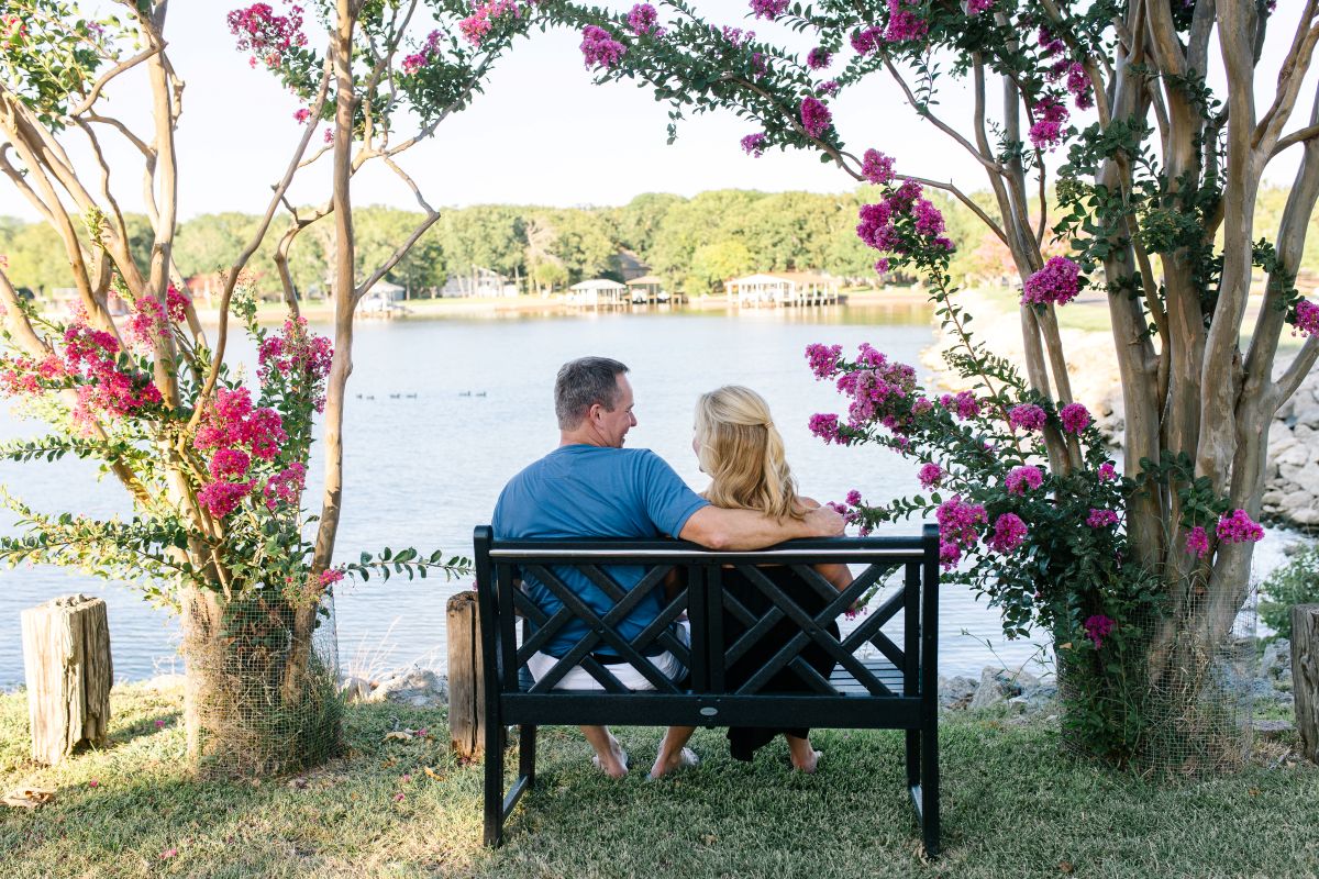 A man and woman sit on a black POLYWOOD Chippendale garden bench between flowering trees by a lake.