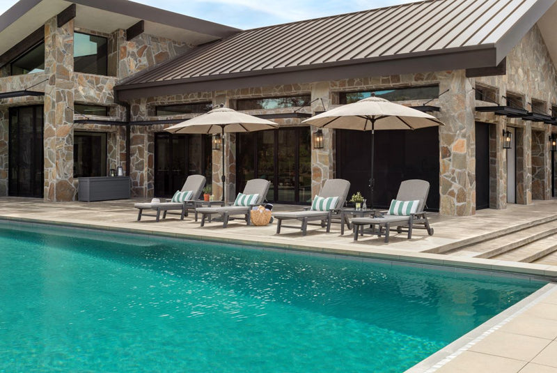 Under umbrellas, a grey POLYWOOD Nautical Chaise 6 Piece Set with Arms and Wheels sits by a flagstone home on a poolside patio.
