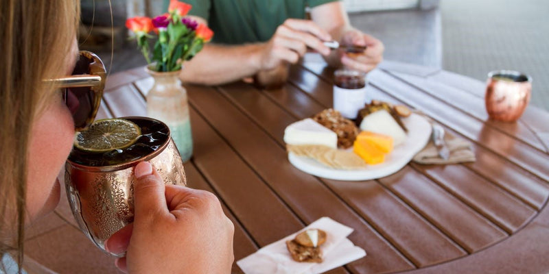Woman enjoys a Moscow Mule on an outdoor patio table with snacks on the table.