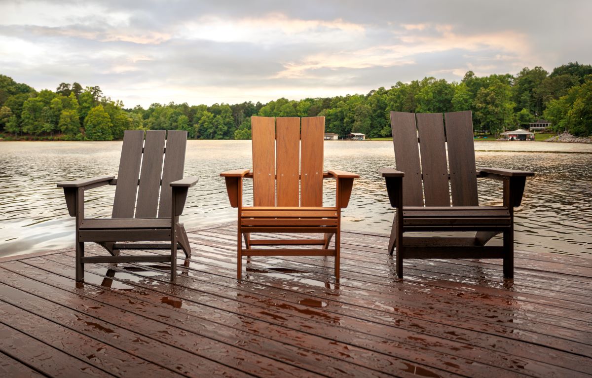 Three brown Adirondack chairs from the POLYWOOD Modern Adirondack Collection sit on a wooden dock on a lake lined with trees.