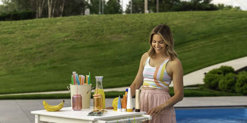 A woman stands by a white POLYWOOD outdoor bar cart holding sunscreen bottles, sunglasses, popsicles, and cold drinks.