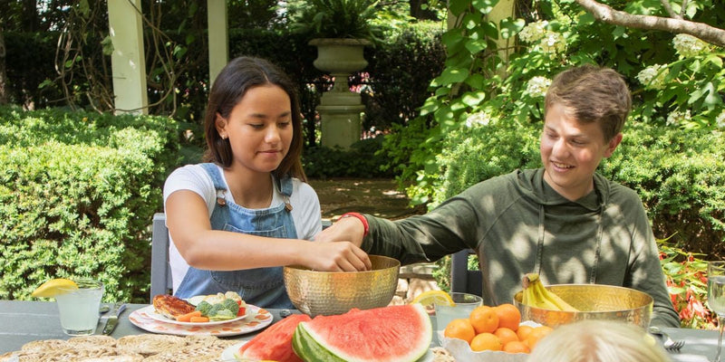 Kids eat lunch at a grey POLYWOOD dining table with plates of grilled food, fruit, and lemonade, surrounded by lush greenery.