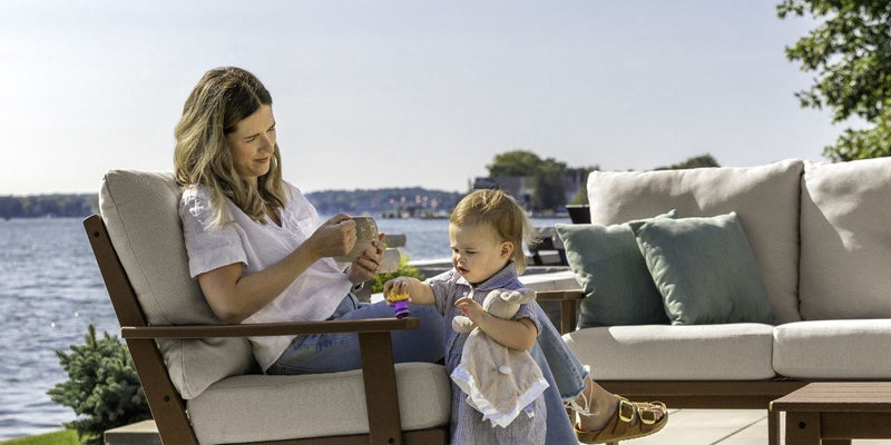 On a lakefront patio, a woman sits in a brown POLYWOOD Prairie Deep Seating Chair with tan cushions; a toddler stands nearby.