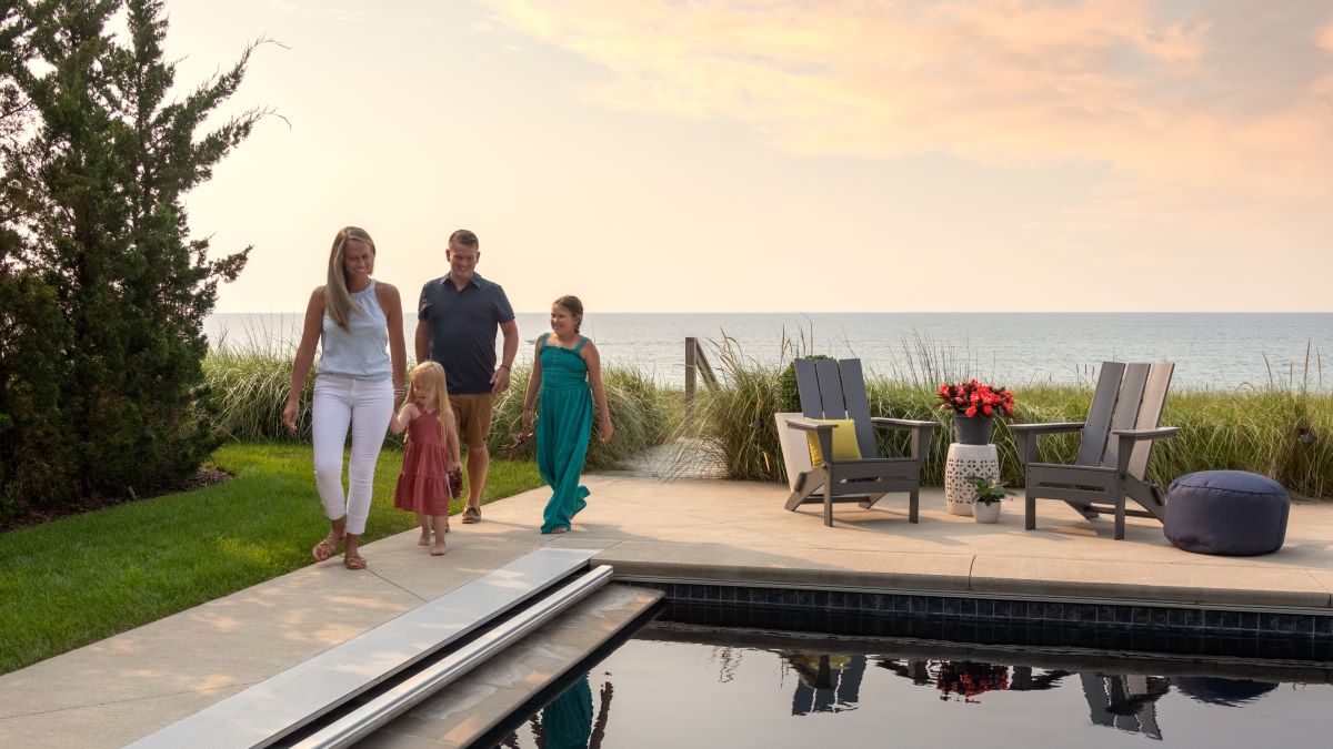 A family walks by grey POLYWOOD Modern Folding Adirondacks on a poolside patio overlooking the ocean.
