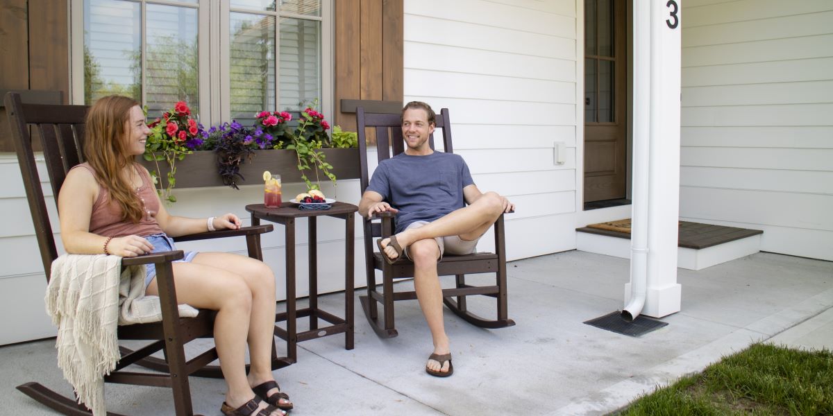 A man and woman sit on a brown POLYWOOD Vineyard 3 Piece Rocking Set on a white home’s porch with shutters and a flower box.