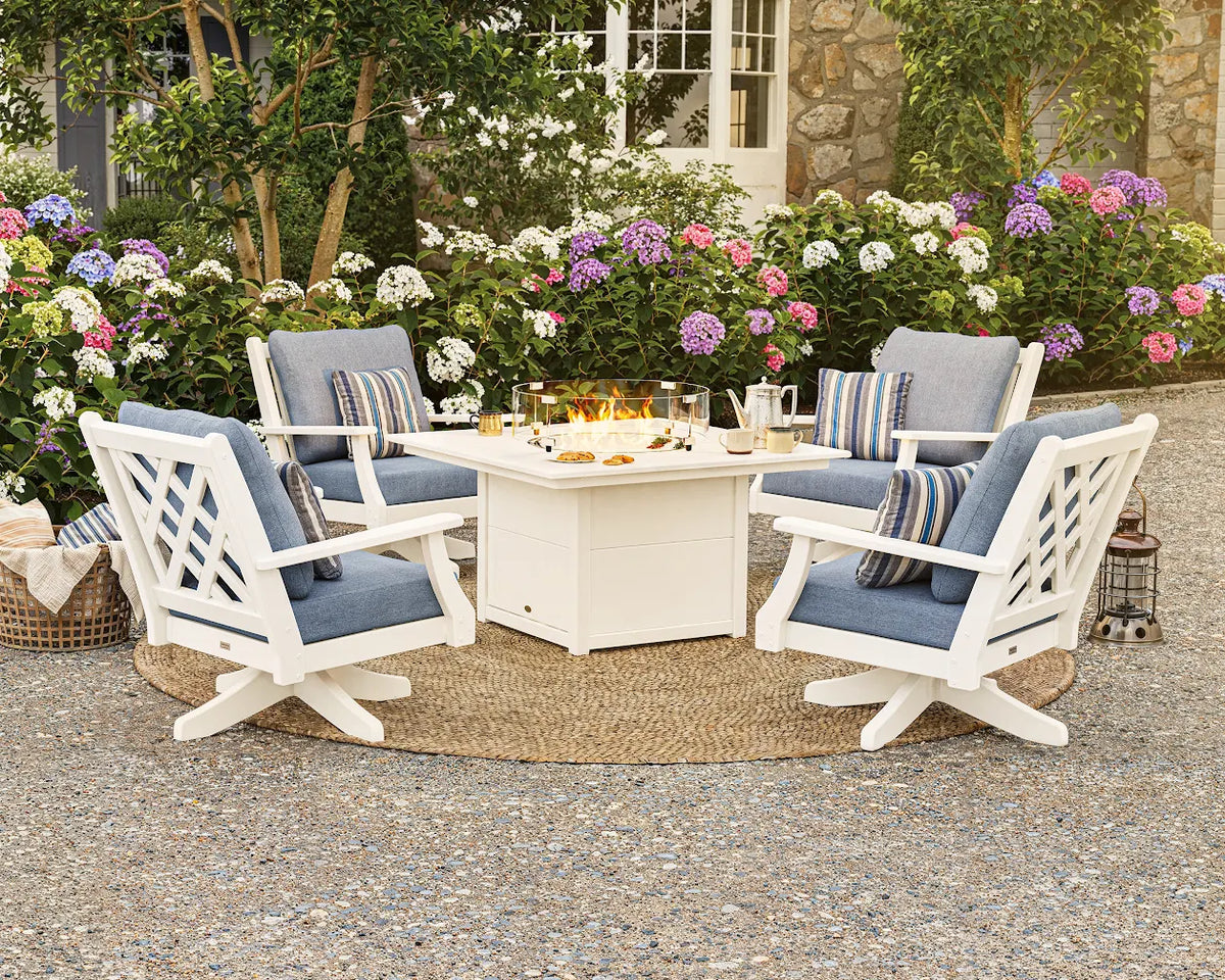 A white POLYWOOD Wovendale 5-Piece Deep Seating Swivel Conversation Set with Fire Pit Table and blue cushions sits on a gravel area near hydrangea bushes.
