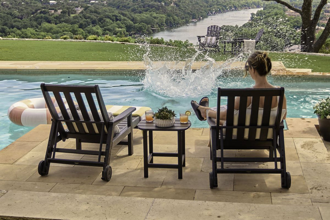 A woman sits in a black POLYWOOD Vineyard 3 Piece Chaise With Arms Set on a poolside patio as someone splashes into the water.