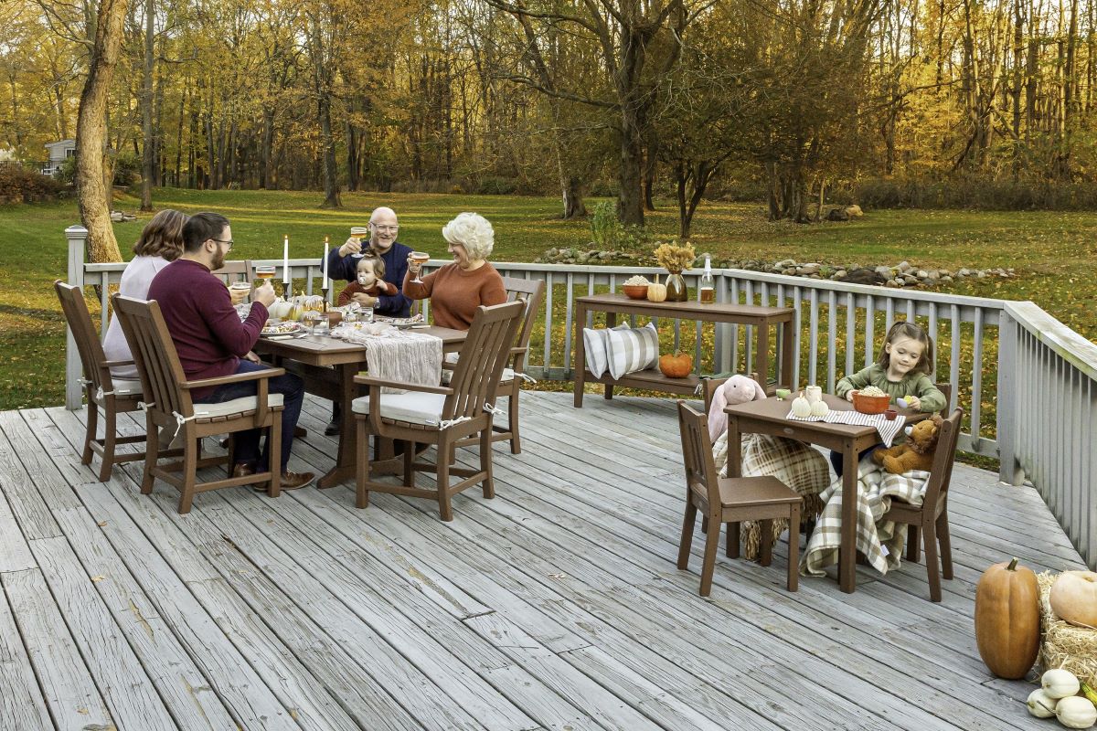 A family gathers for a meal at assorted brown POLYWOOD dining sets on a wooden deck with fall decor by a lawn and tall trees.