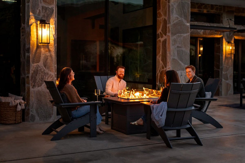 People sip wine in black POLYWOOD Modern Curveback Adirondack Chairs around a Rectangle 34 by 46 Inch Fire Pit Table on a patio.