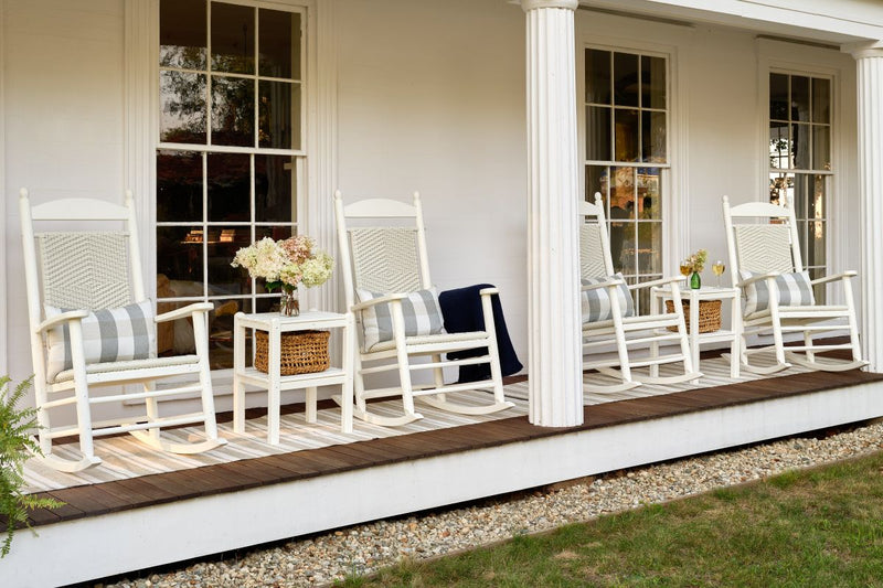 Jefferson Woven Rocking Chairs and Two Shelf Side Tables sit on a wooden covered porch before a white house.