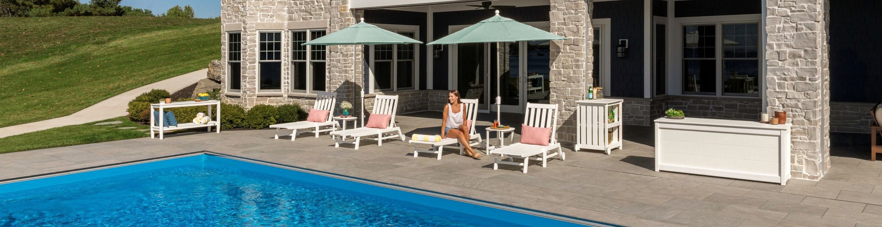 A woman sits on a POLYWOOD Vineyard 3-Piece Chaise with Wheels Set on a poolside patio by a stone house.
